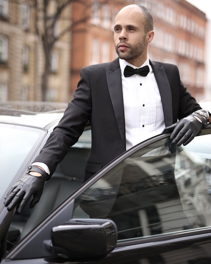 Well-dressed man in a tuxedo standing confidently by his car in an urban setting.